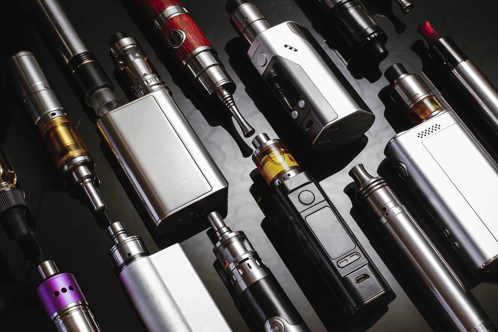 Close-up overhead shot of multiple vaping devices and box mods on a black surface.